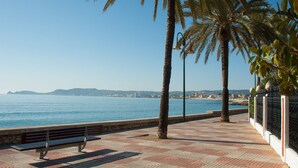 Une promenade en bord de mer, agrémentée d’un banc, de palmiers et offrant une vue dégagée sur la mer et les collines lointaines.
