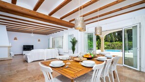 A spacious dining area with a wooden table set for four, surrounded by white chairs. A large window offers a view of a swimming pool and greenery. - Villa Alma in El Tosalet (Javea)