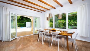 A dining area with a wooden table and white chairs, large windows with a view of greenery, and a glass door leading outside. - Villa Alma in El Tosalet (Javea)