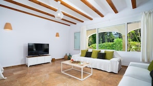 A modern living room with a flat-screen TV, white sofas, a coffee table, and large windows offering a view of greenery. - Villa Alma in El Tosalet (Javea)