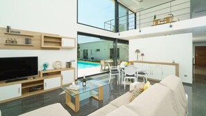 A modern living room with a flat-screen TV, glass coffee table, and a sofa. There is a dining area with a table and chairs, and a view of a swimming pool through large windows. - Villa Vera in Javea With 4 Bedrooms and 2 Bathrooms (Javea)