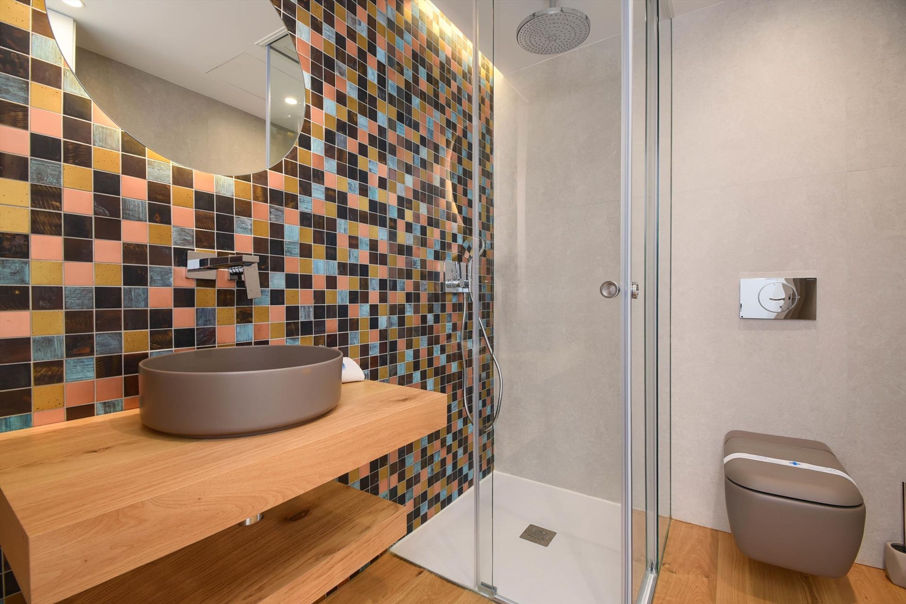 A modern bathroom with a tiled backsplash, a round sink, a glass shower enclosure, and a toilet.