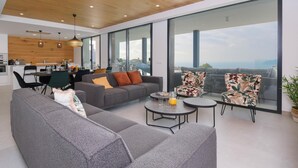 A modern living space with a grey sofa, a dining area, and large windows offering a city view. - Villa Madi in Cumbre del Sol (Benitachell)
