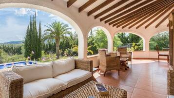 Un área de estar al aire libre techada con sofá y sillones, con vista a una piscina y abundante vegetación.