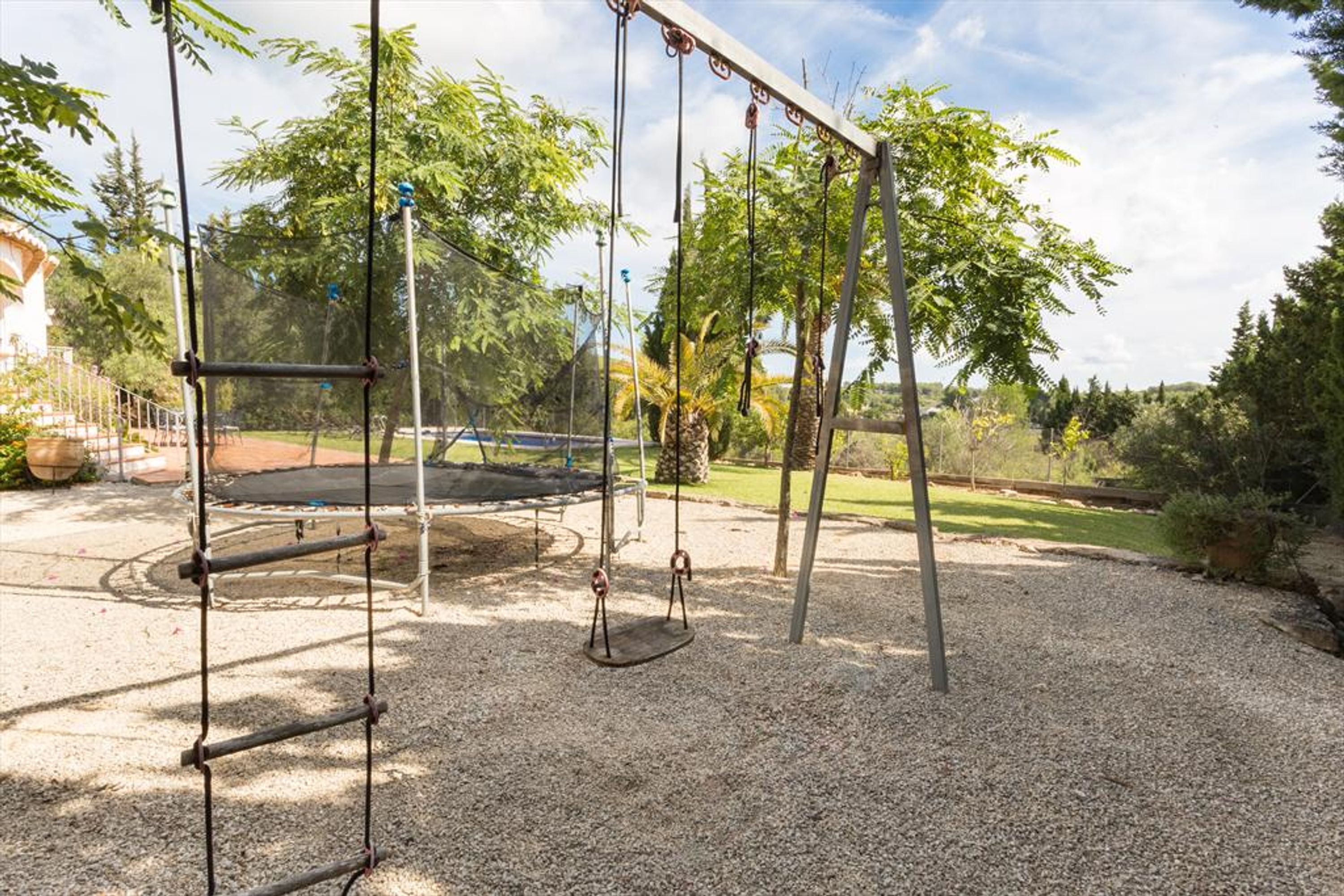 A playground with swings and a trampoline, surrounded by gravel and trees.