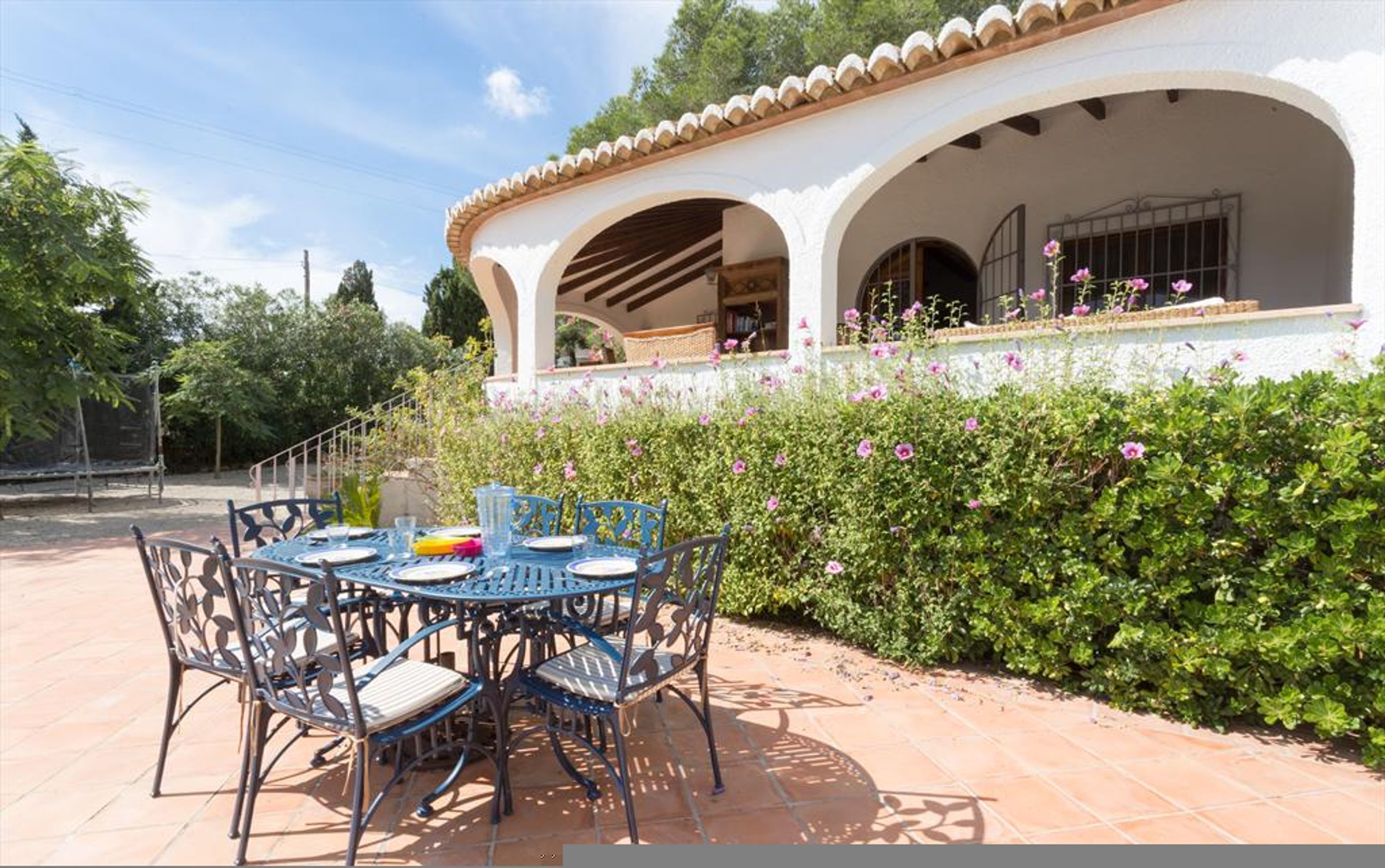 Un patio al aire libre con una mesa redonda y sillas, un edificio blanco con arcos y una cobertura vegetal con flores rosadas.