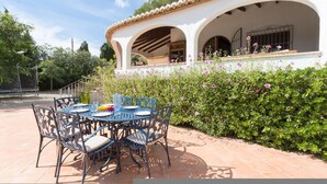 An outdoor patio with a round table and chairs, a white building with arches, and a hedge with pink flowers. - Villa Yuca in La Mandarina (Javea)