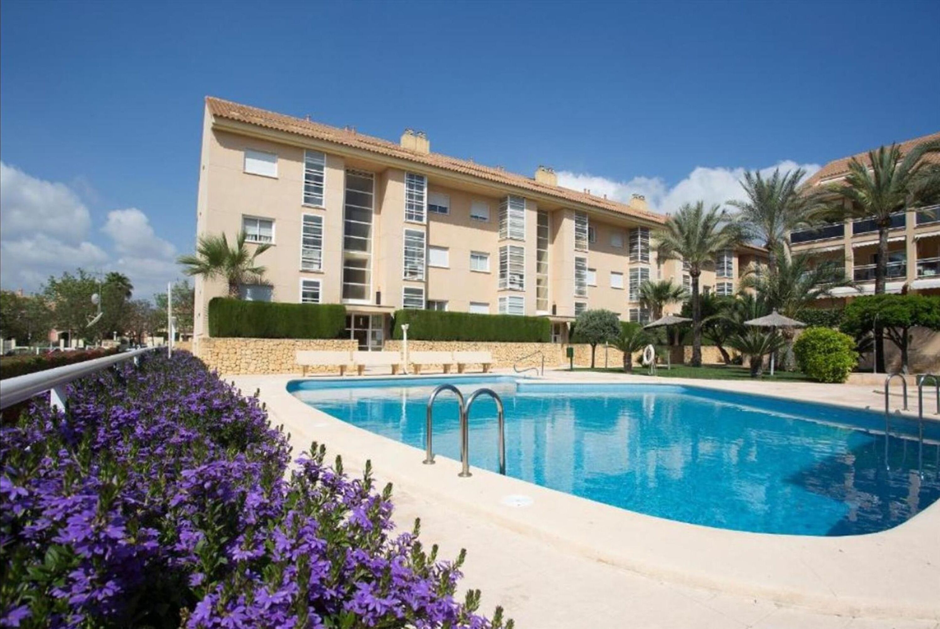 A hotel with a swimming pool, a well-maintained garden, and a clear blue sky.