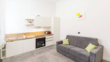 A modern kitchen with white cabinets, a grey sofa with green cushions, and a small plant on a shelf.