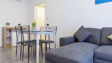 Una sala de estar moderna con un sofá gris, una mesa de comedor con sillas y una pequeña planta sobre una mesita de madera.