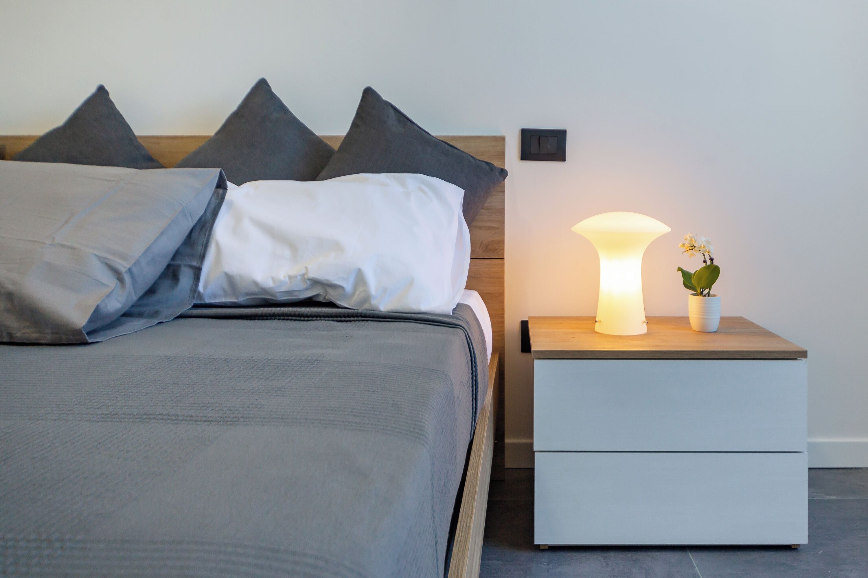 Ein ordentlich bezogenes Bett grau, ein Nachttisch mit Lampe und Topfpflanze, und ein Lichtschalter an der Wand.