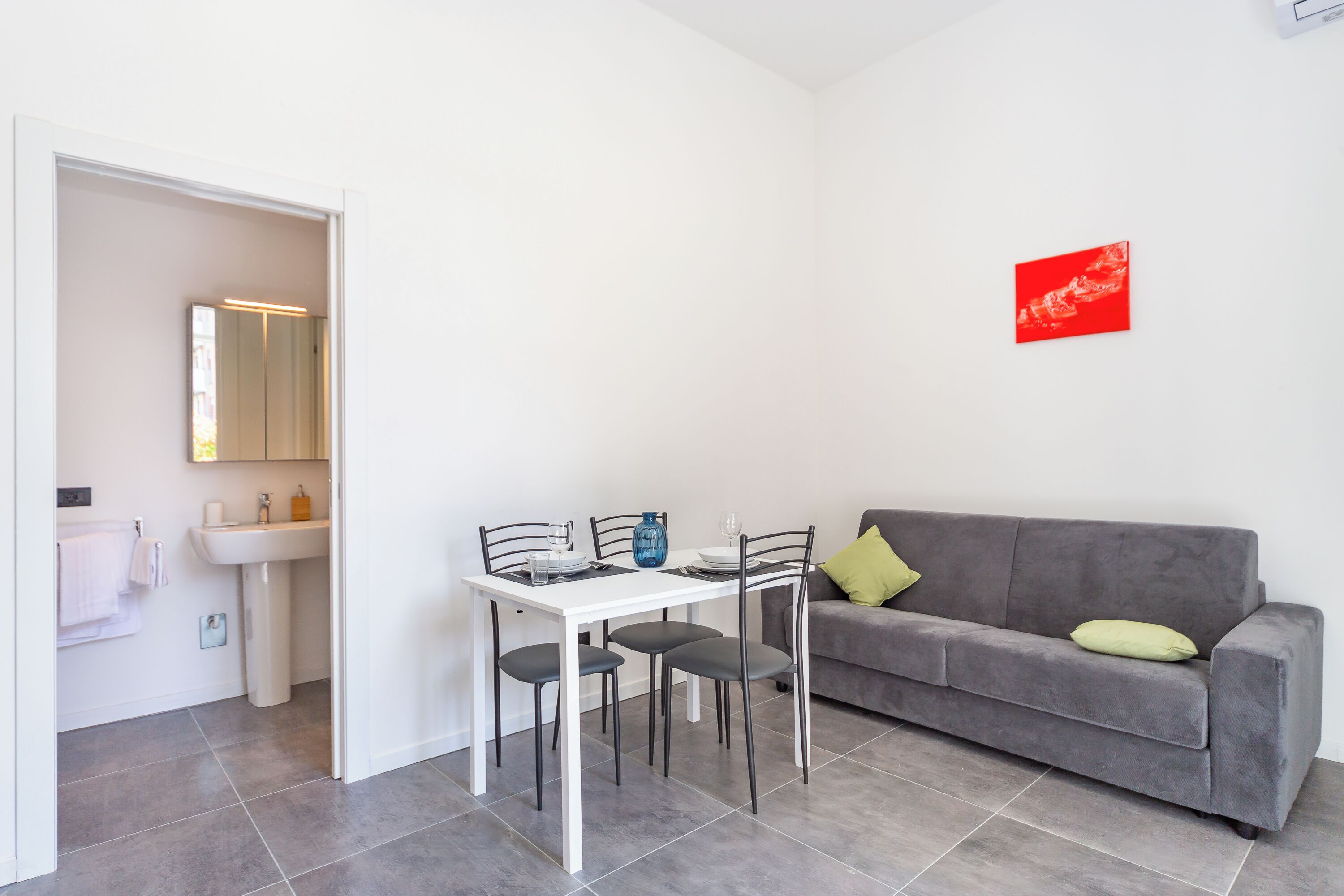 A modern living space with a dining table, four chairs, a grey sofa with a green pillow, and a red abstract painting on the wall.