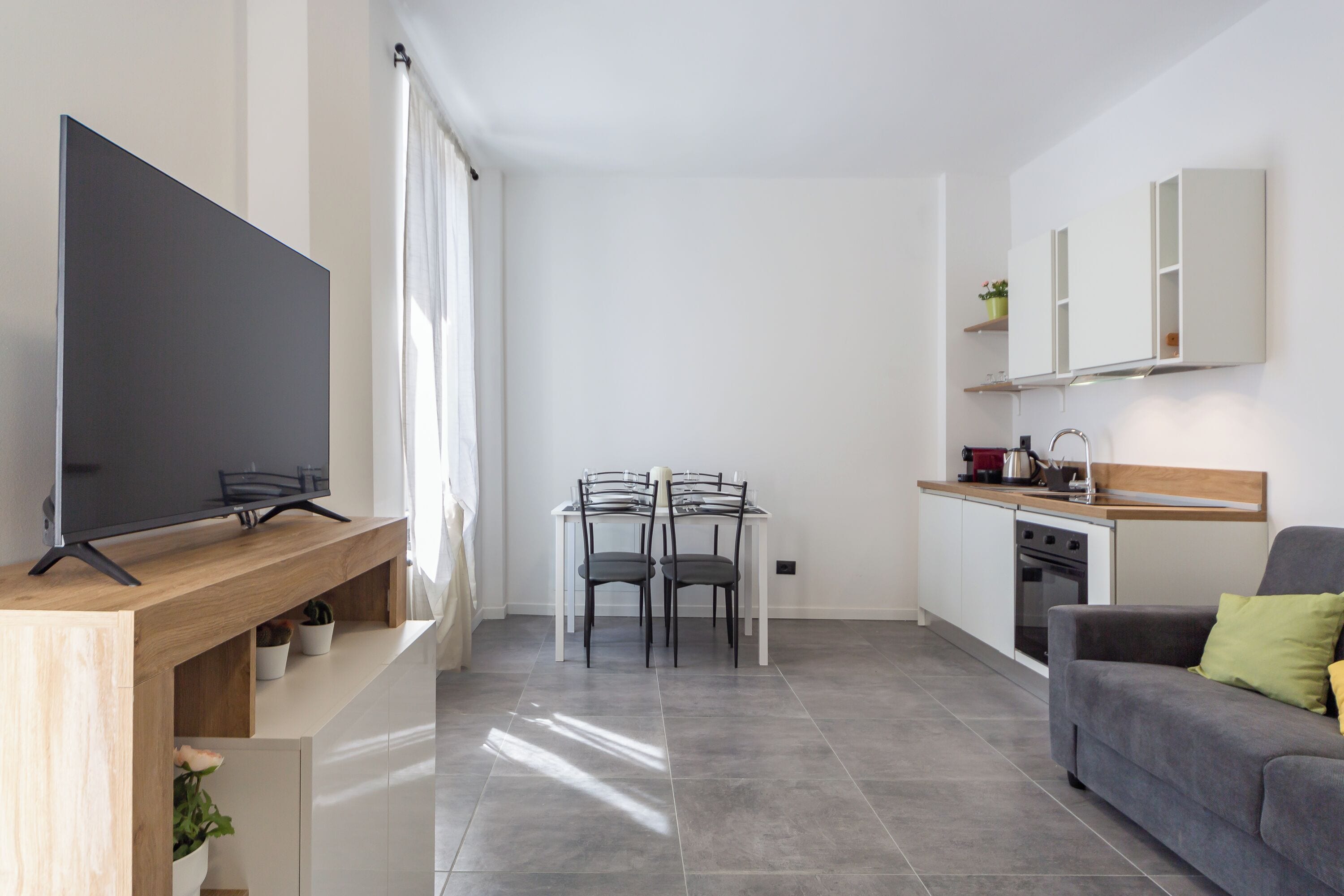 Una sala de estar moderna con un televisor de pantalla plana sobre un mueble de madera, un sofá gris con cojines, una zona de comedor con mesa y sillas, y una cocina con armarios blancos y un horno empotrado.