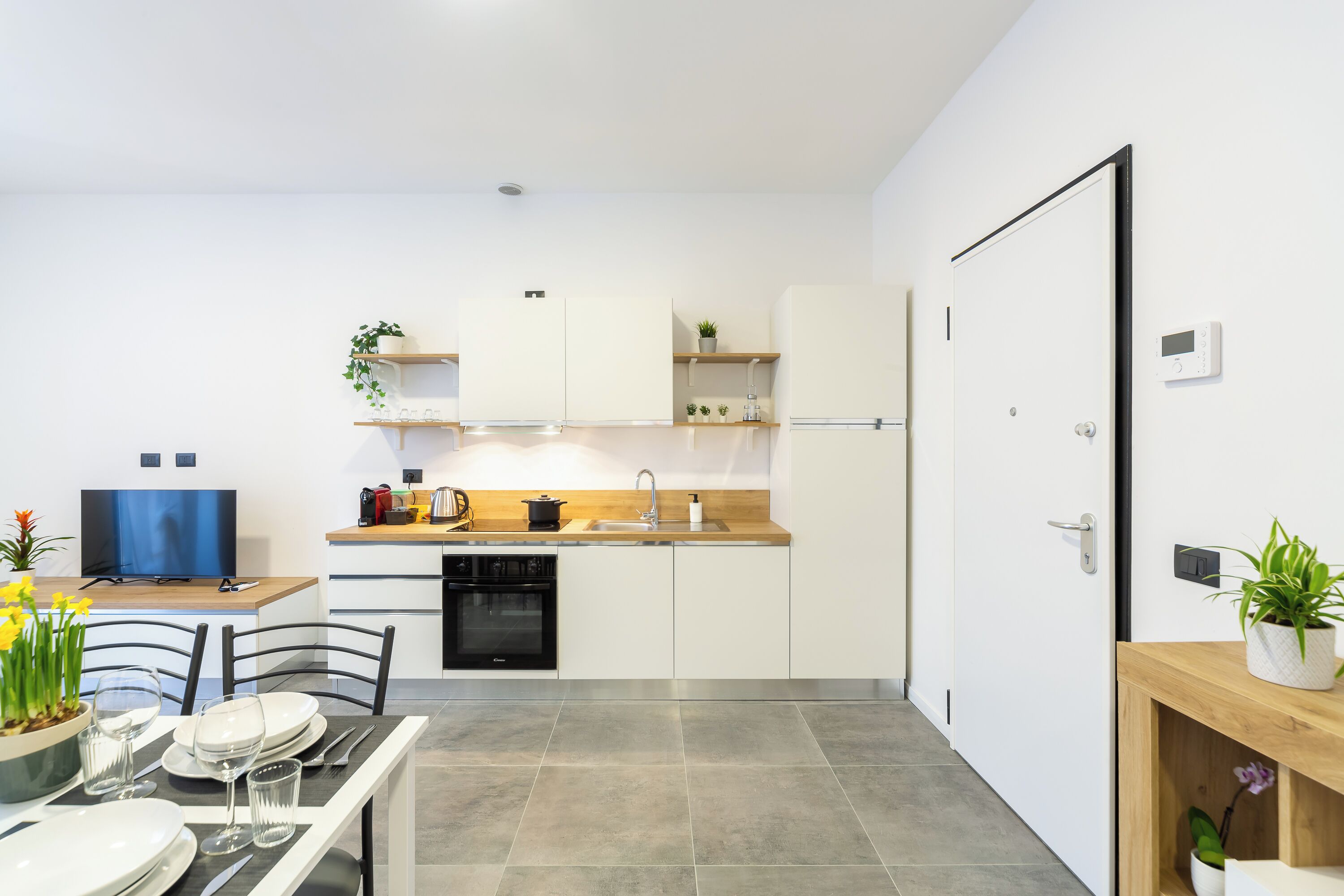 Une cuisine moderne avec des armoires blanches, un plan de travail en bois et un lave-vaisselle encastré. On y trouve un coin repas avec une table pour deux personnes et un téléviseur posé sur un meuble.