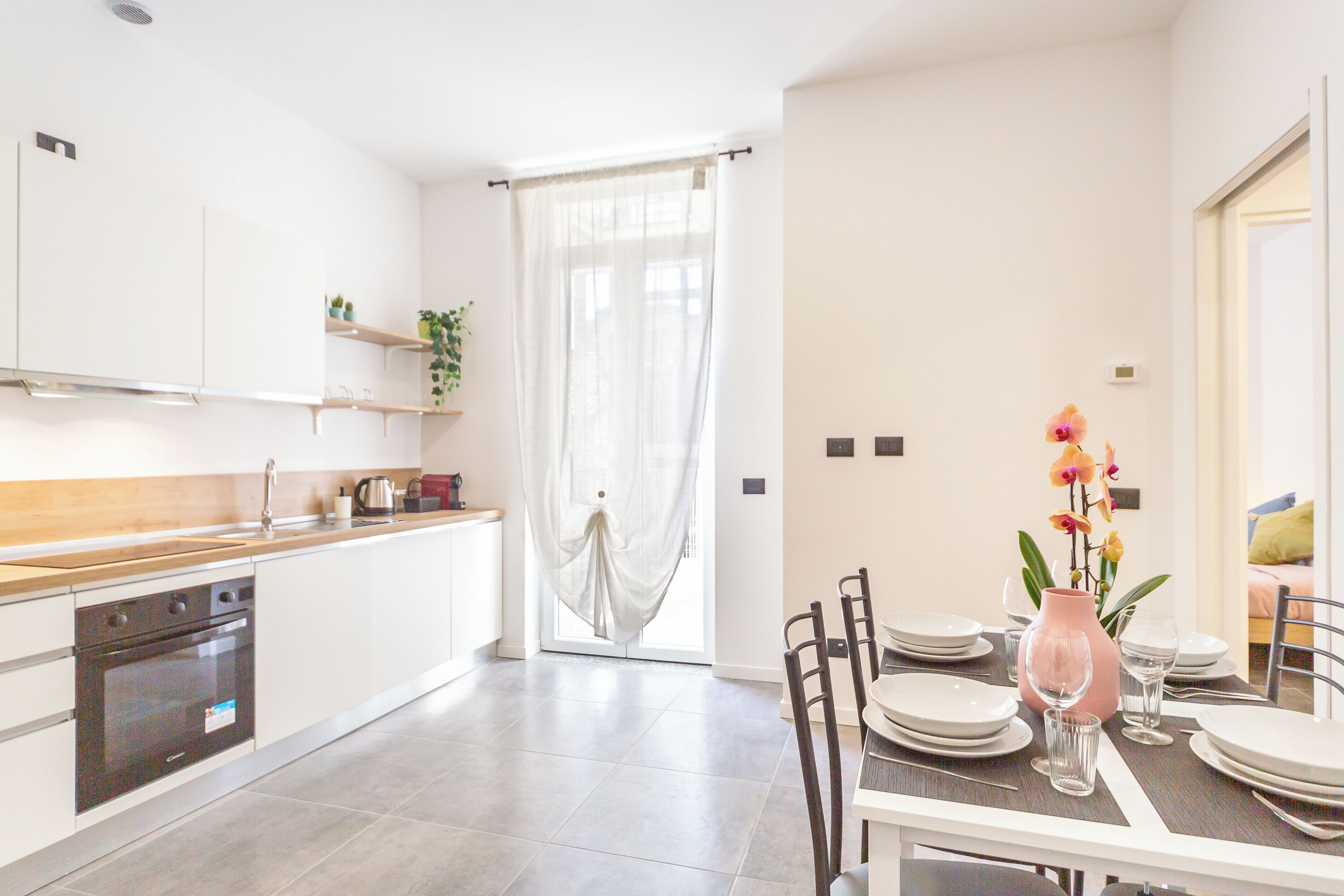 A modern kitchen with white cabinets, a wooden countertop, and a built-in oven. There is a dining area with a table set for four, a pink vase with flowers, and a window with sheer curtains.