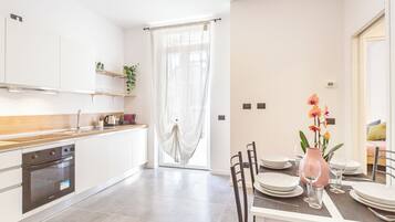 A modern kitchen with white cabinets, a wooden countertop, and a built-in oven. There is a dining area with a table set for four, a pink vase with flowers, and a window with sheer curtains.