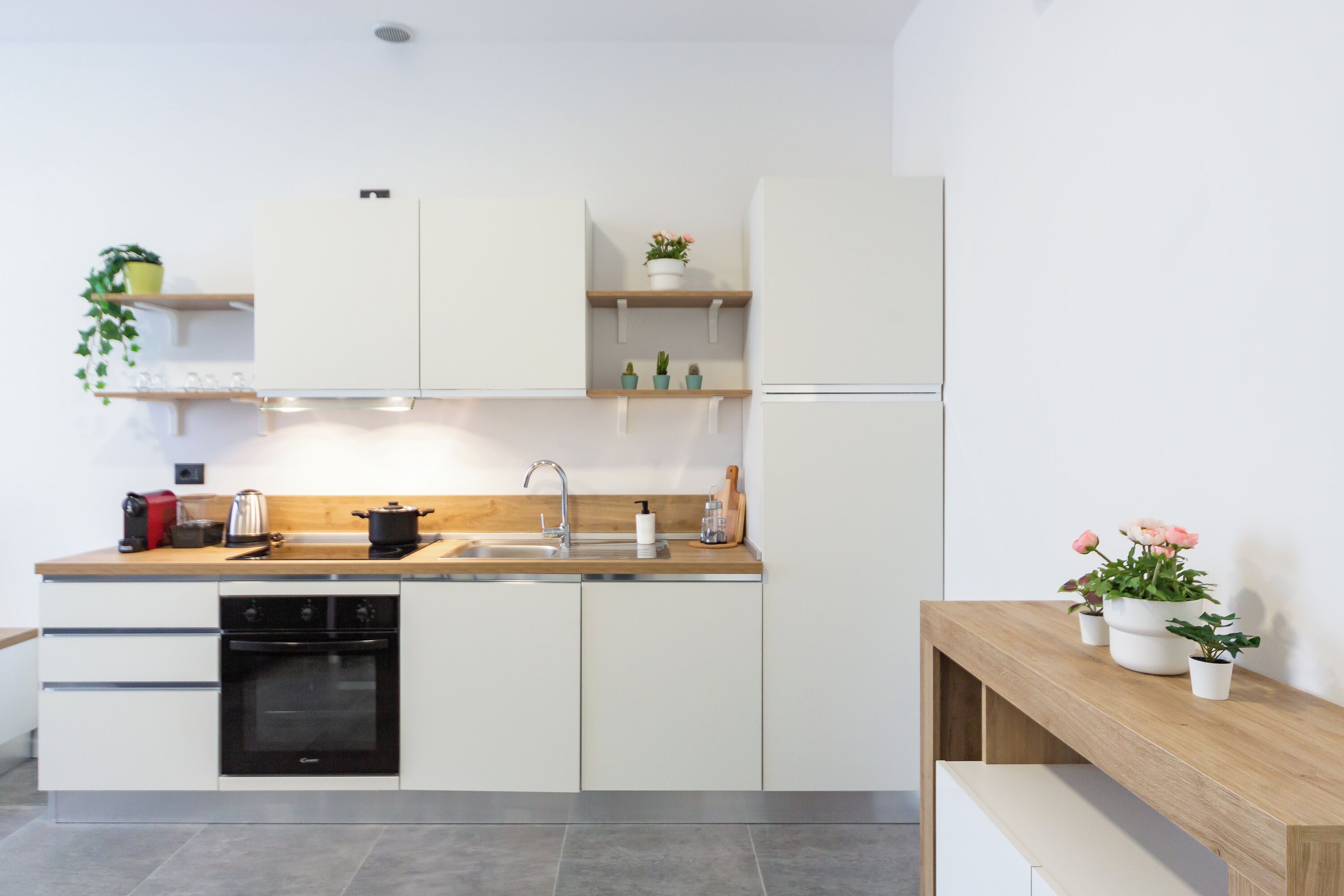 A modern kitchen with white cabinets, a wooden countertop, a built-in oven, and a sink. There are potted plants on shelves and a wooden bench.