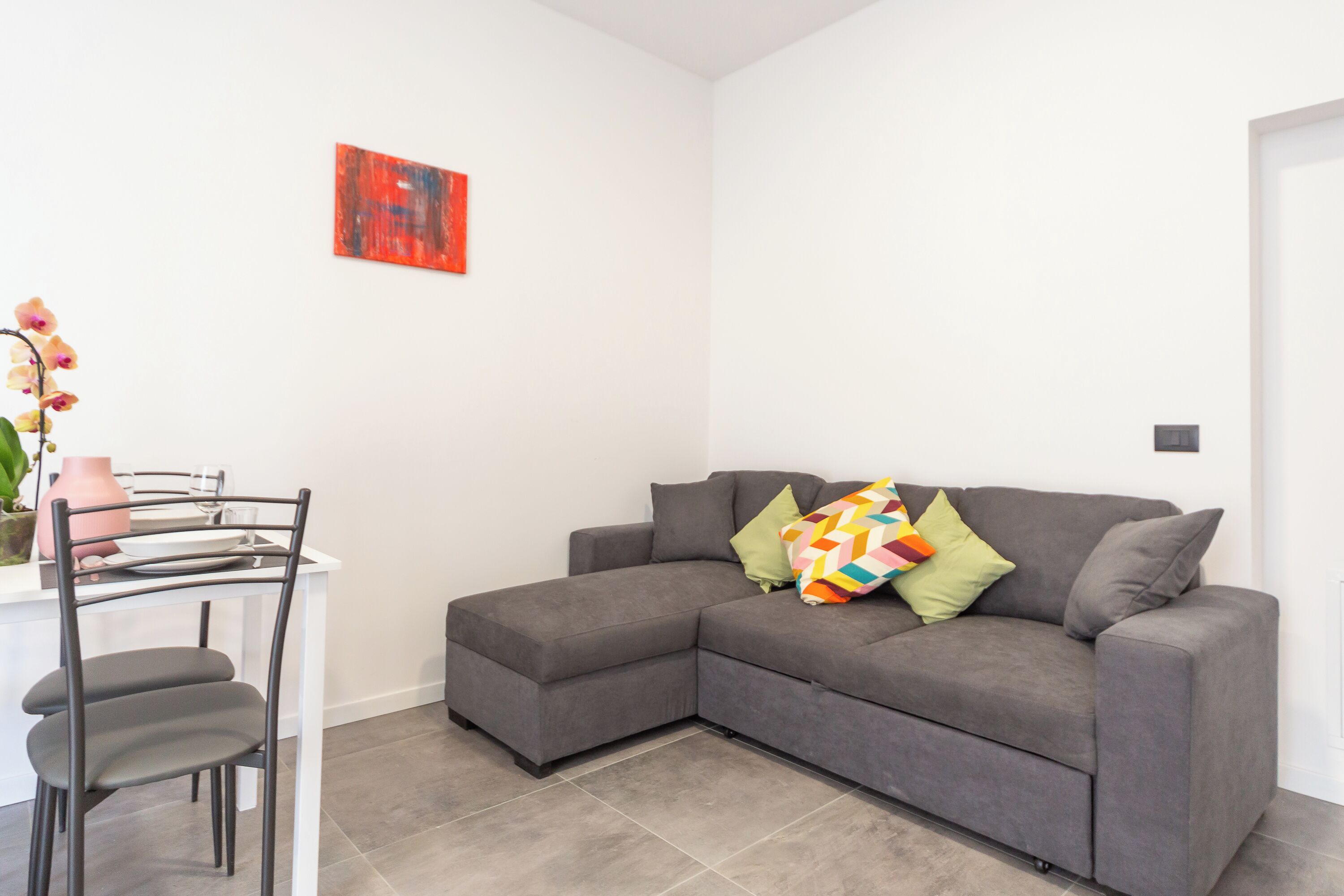 Ein modernes Wohnzimmer mit einem grauen Ecksofa, einem weißen Esstisch mit Stühlen und einer einzelnen Orchideenpflanze.