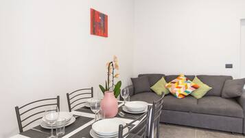 Un coin repas avec une table dressée pour quatre personnes, des chaises, un vase avec des fleurs et un canapé gris agrémenté de coussins colorés.