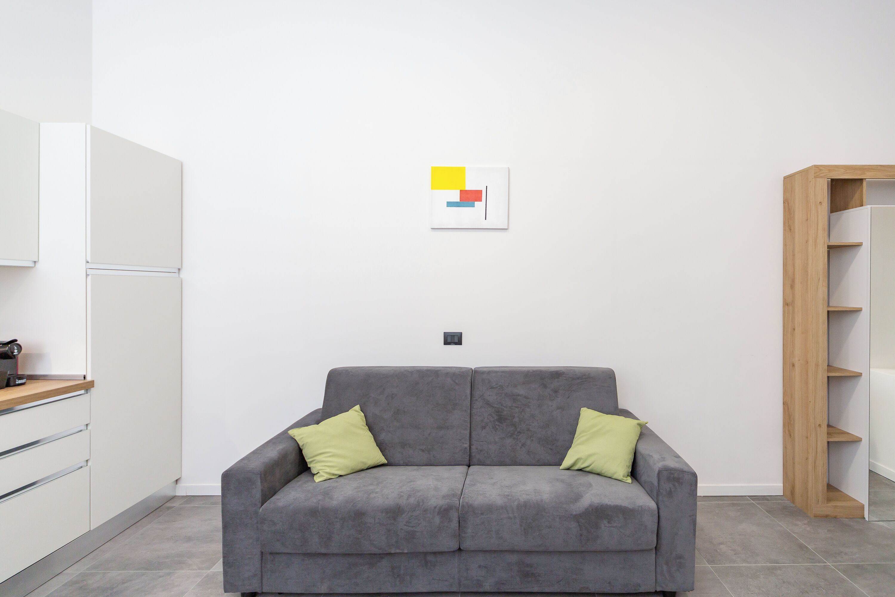 Ein modernes Wohnzimmer mit einem grauen Sofa, zwei grünen Kissen, einer Holzkommode und einer weißen Wand mit einem geometrischen Kunstwerk.
