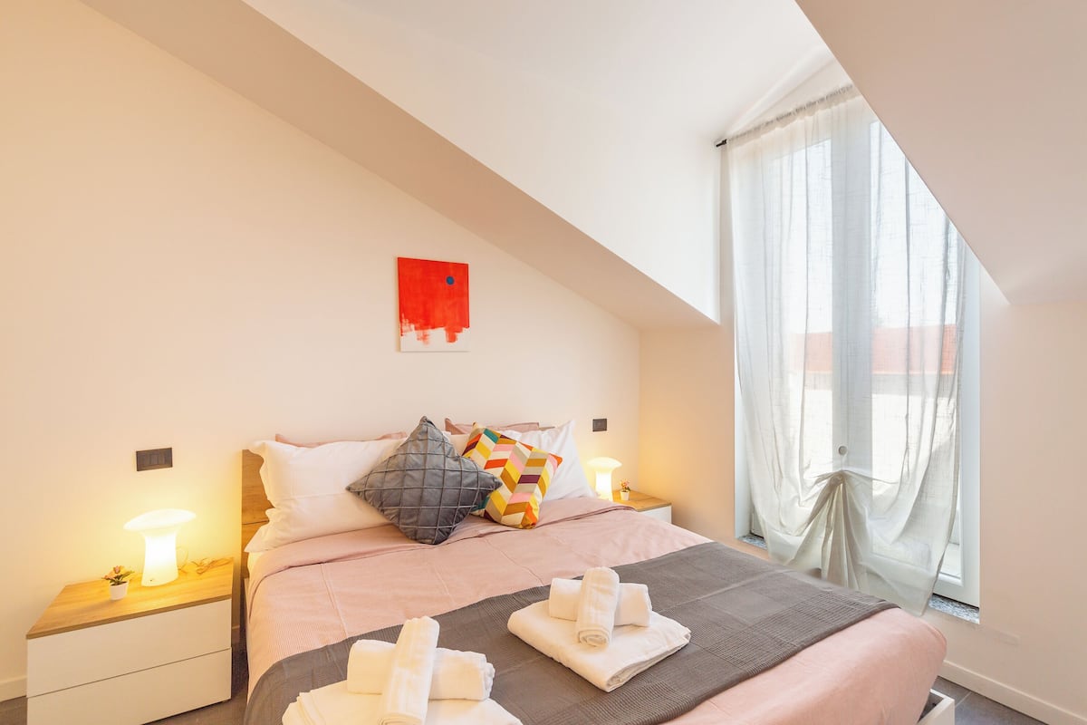 A bedroom with a bed, pillows, a bedside table with a lamp, and a window with curtains.