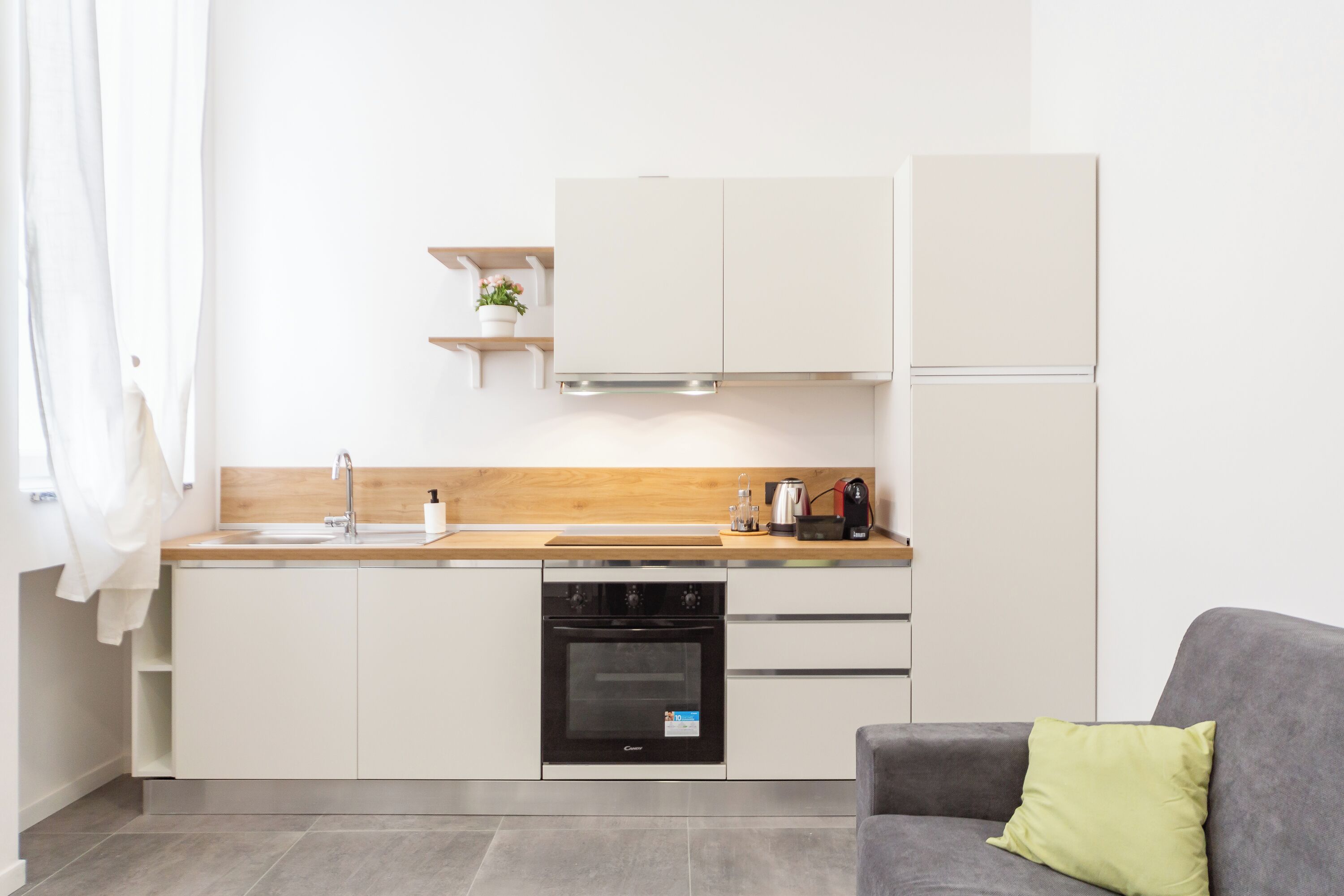 Eine moderne Küche mit weißen Schränken, einer Holz Arbeitsplatte, einem Einbauofen und einem grauen Sofa mit einem grünen Kissen.