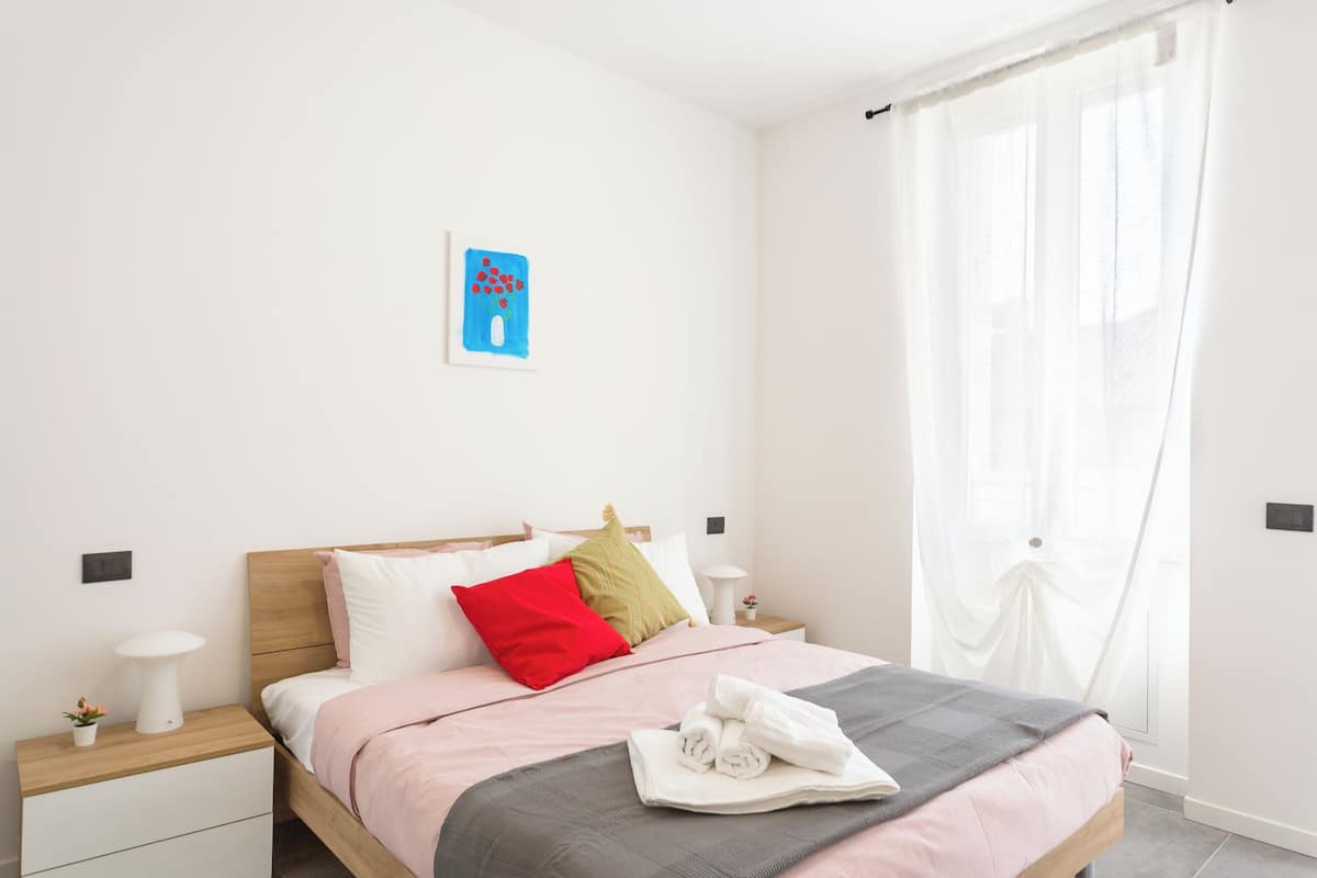A neatly made bed with a pink and gray blanket, a red pillow, and a white towel on the side. There is a small table with a lamp and a plant on the left.