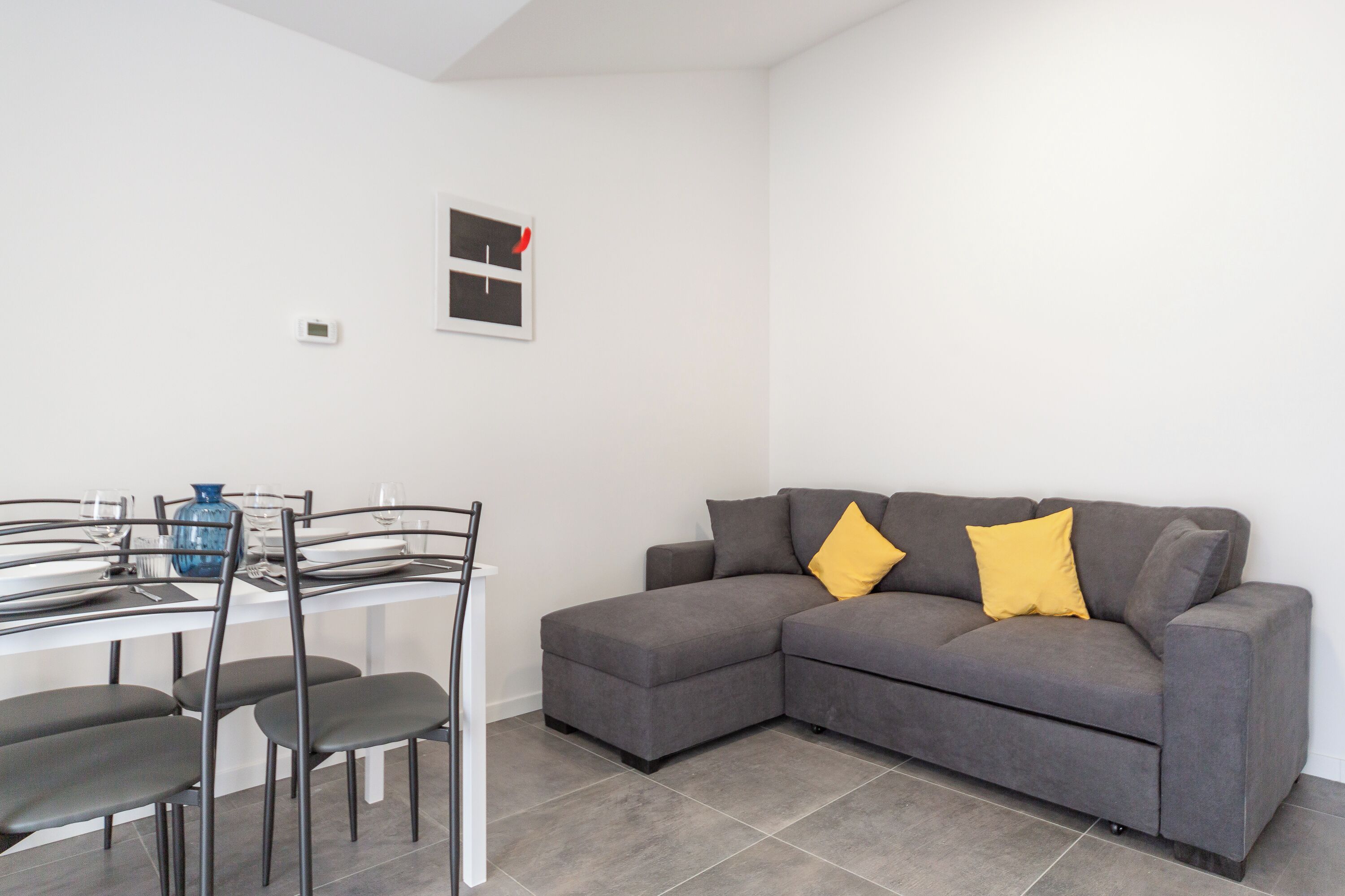 A modern living room with a grey sectional sofa, a dining table with chairs, and a blue vase.