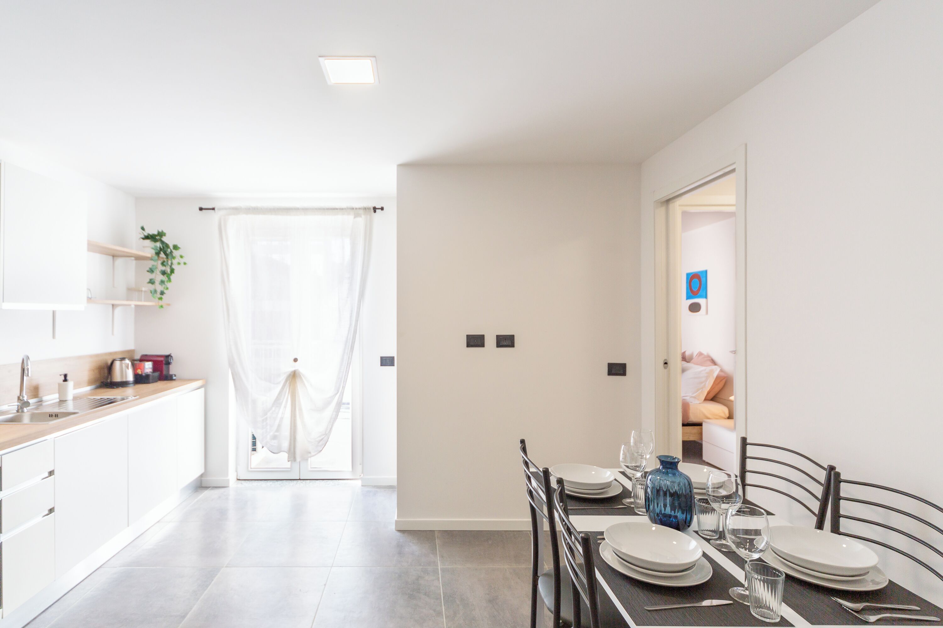 A modern kitchen with white cabinets, a sink, and a plant. A dining area with a table set for two is visible.