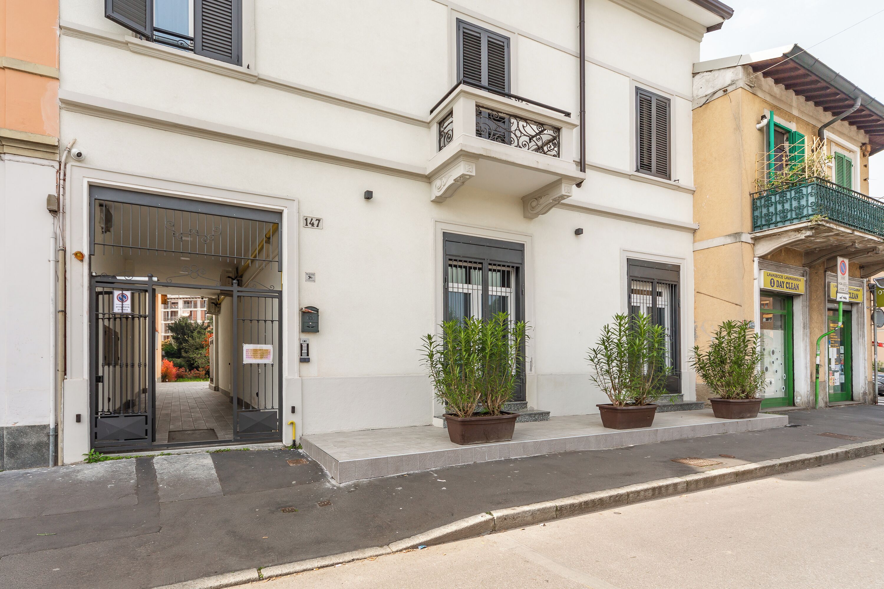 Un immeuble doté d’une entrée fermée par un portail, d’un balcon orné d’une balustrade en fer forgé et de plantes en pot disposées sur le trottoir.
