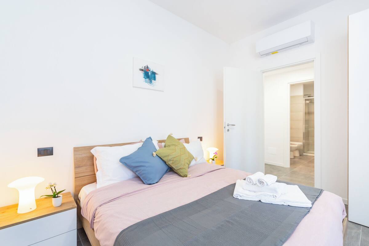 A modern hotel room with a neatly made bed, a bedside table with a lamp and a plant, and a door leading to the bathroom.