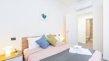 Une chambre d’hôtel moderne avec un lit bien fait, une table de chevet avec une lampe et une plante, et une porte donnant accès à la salle de bain.