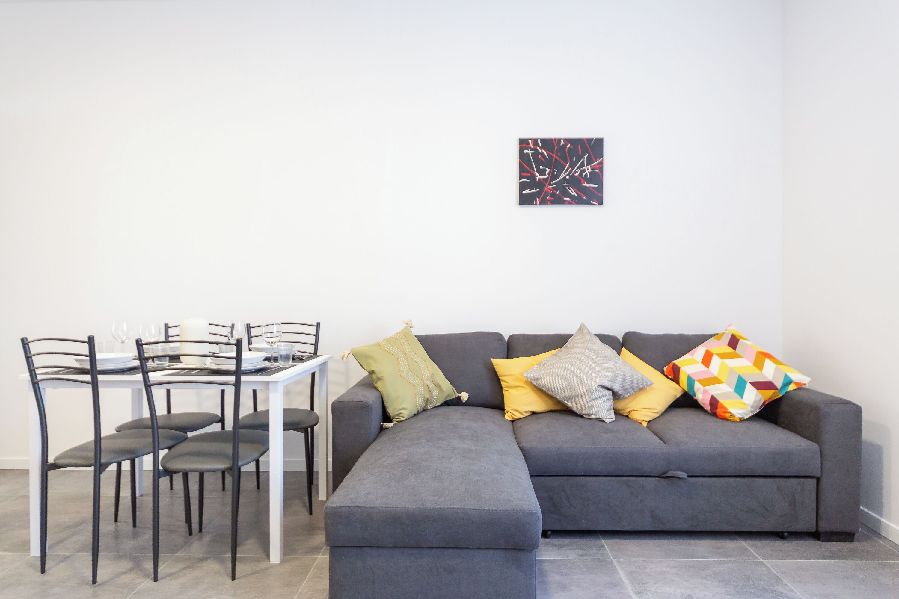 A modern living room with a grey sectional sofa, a dining table with chairs, and a small abstract painting on the wall.