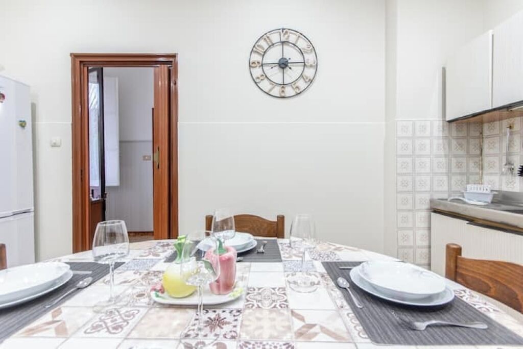 Een eethoek met een tafel voor vier personen, een wandklok en een keuken op de achtergrond.