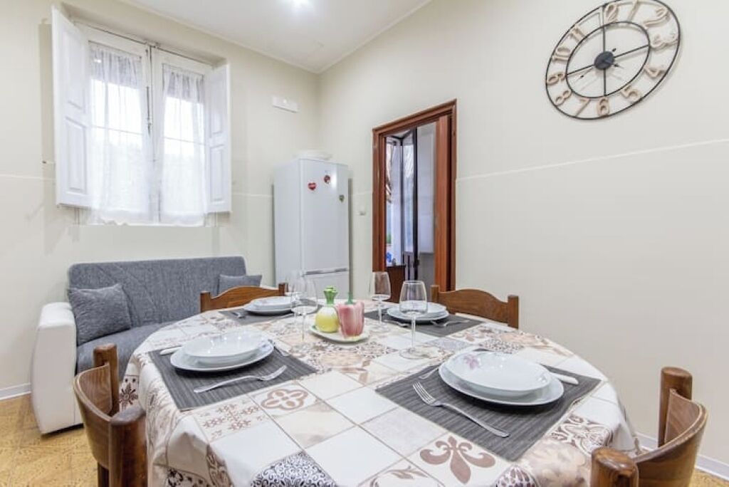 Un coin repas avec une table ronde dressée pour quatre personnes, une horloge au mur et une fenêtre avec des rideaux.
