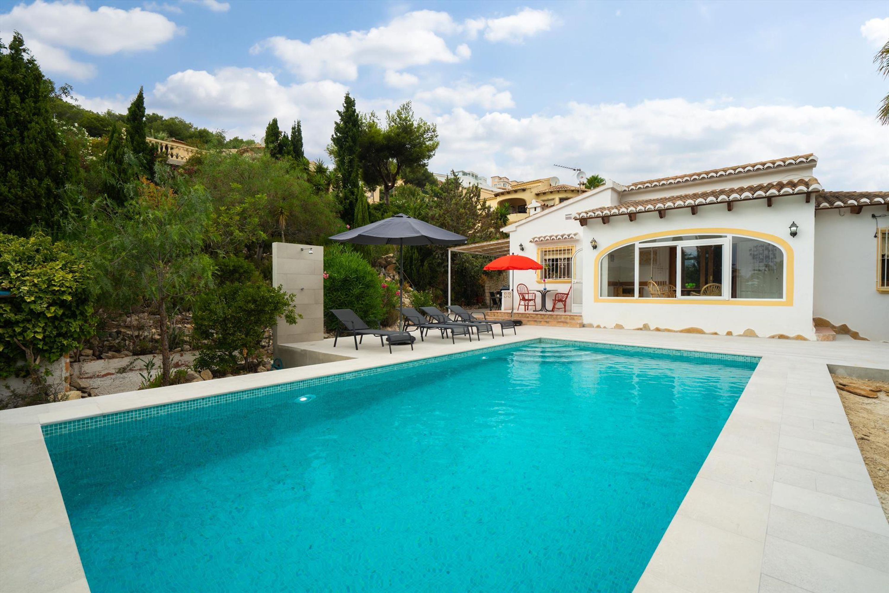 Piscina con sillones de descanso y sombrillas, rodeada de vegetación y una colina.