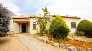 A two-story house with a tiled roof, a front entrance with a door, and a small porch. There are various plants and shrubs in the front yard, and a pathway leading to the entrance. - Fanila in J vea With 4 Bedrooms and 3 Bathrooms (Javea)