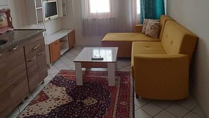 A living room with a yellow sofa, a coffee table, and a kitchen area with cabinets and a TV. - Karya Konaklama (Antalya)