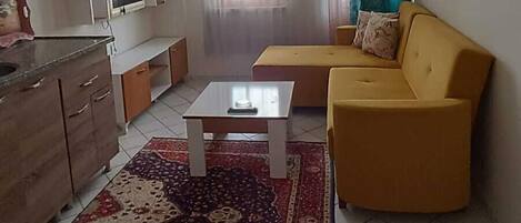 A living room with a yellow sofa, a coffee table, and a kitchen area with cabinets and a TV.