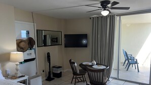 A hotel room with a bed, a desk with a lamp, a chair, a table with a vase, a ceiling fan, a TV, and a sliding glass door to a balcony. - Coastal Tides Ocean View Studio (Daytona Beach)