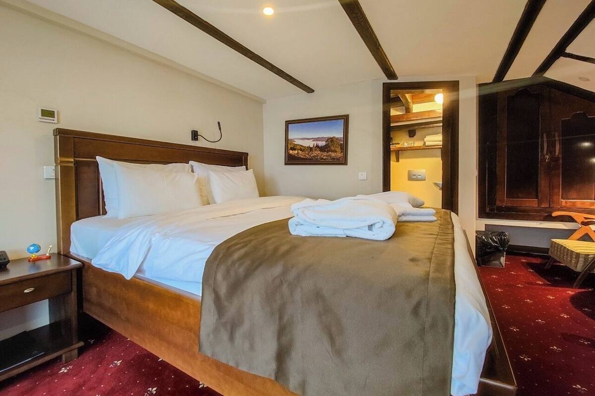 Habitación de hotel con una cama grande, una mesita de noche, un armario de madera y un televisor montado en la pared, además de una alfombra roja.