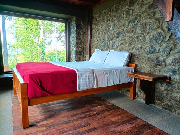 A wooden bed with a red blanket and white sheets, set against a stone wall with a window overlooking greenery.