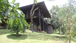 A rustic wooden house with a balcony, surrounded by lush greenery and a well-maintained lawn. -  Entire Property:1BHK Cottage with Attic,Kitchen & Sunset View Deck |Near Munnar (Kambilikandam)