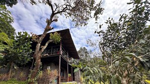 A rustic house with a balcony, surrounded by lush greenery and a clear sky. -  Entire Property:1BHK Cottage with Attic,Kitchen & Sunset View Deck |Near Munnar (Kambilikandam)
