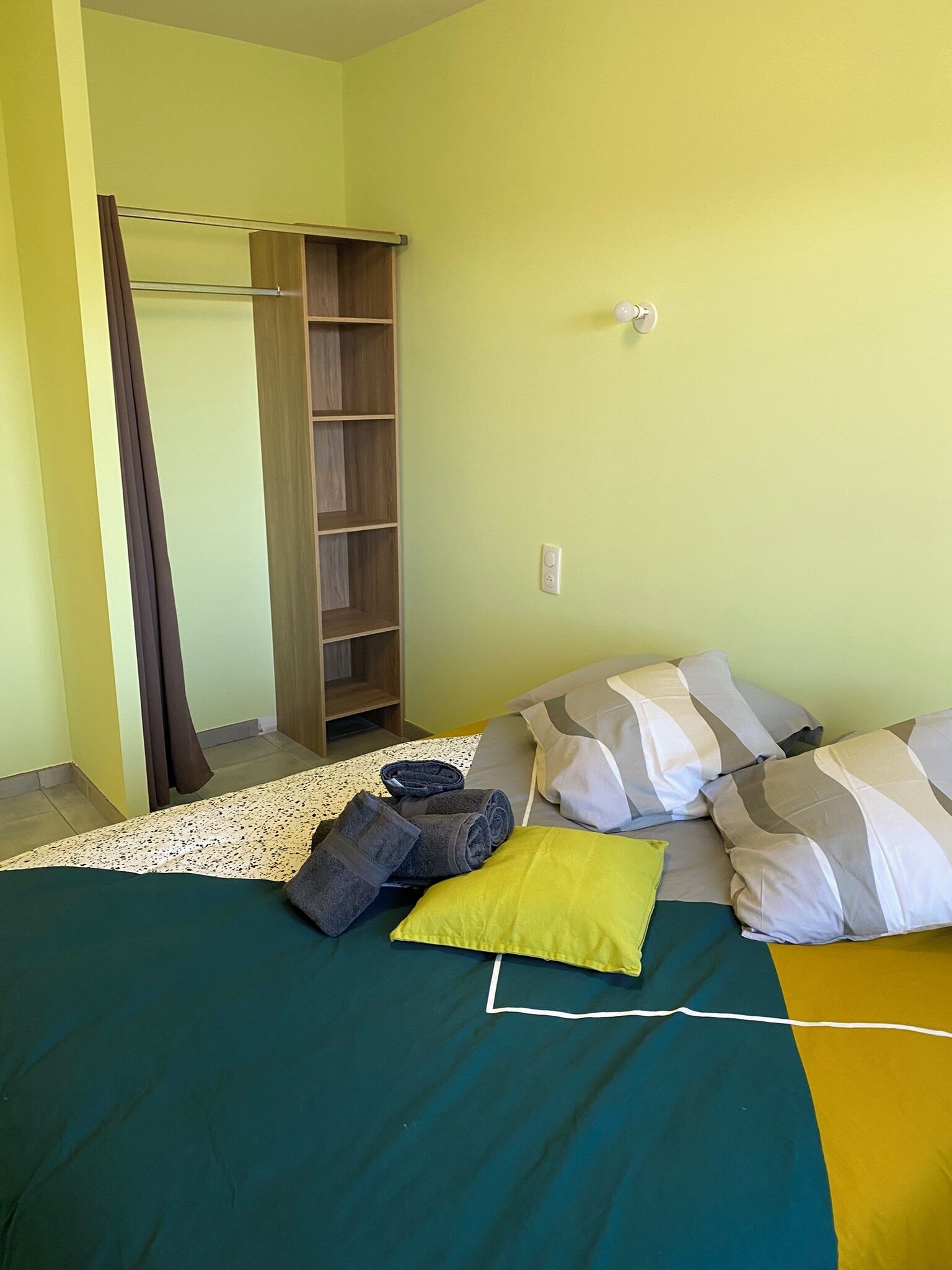 Um quarto com uma cama, almofadas, uma toalha, um guarda-roupa de madeira e um interruptor de luz na parede.