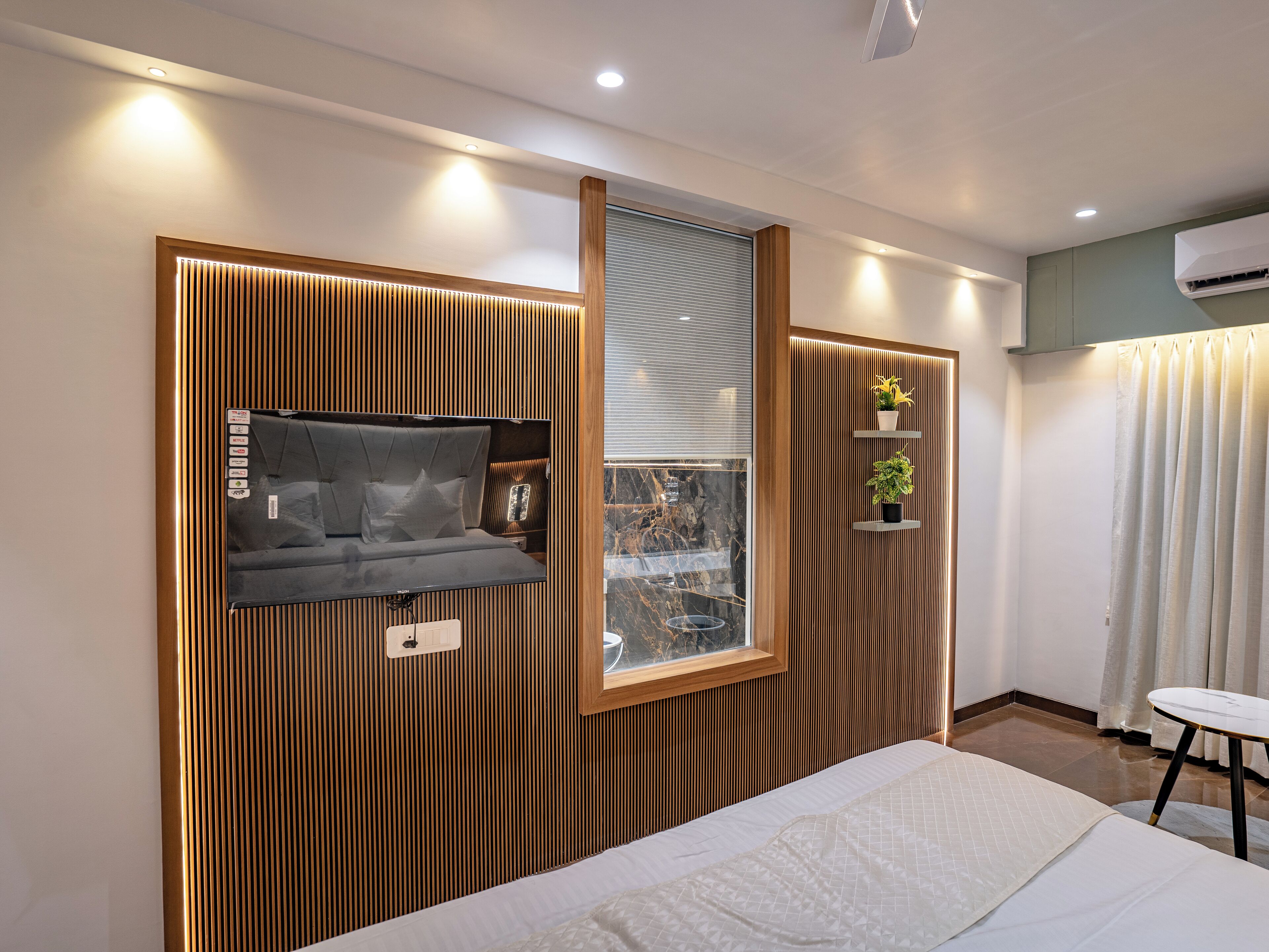 A modern bedroom with a bed, a TV mounted on a wooden panel wall, a window with blinds, and a small shelf with plants.