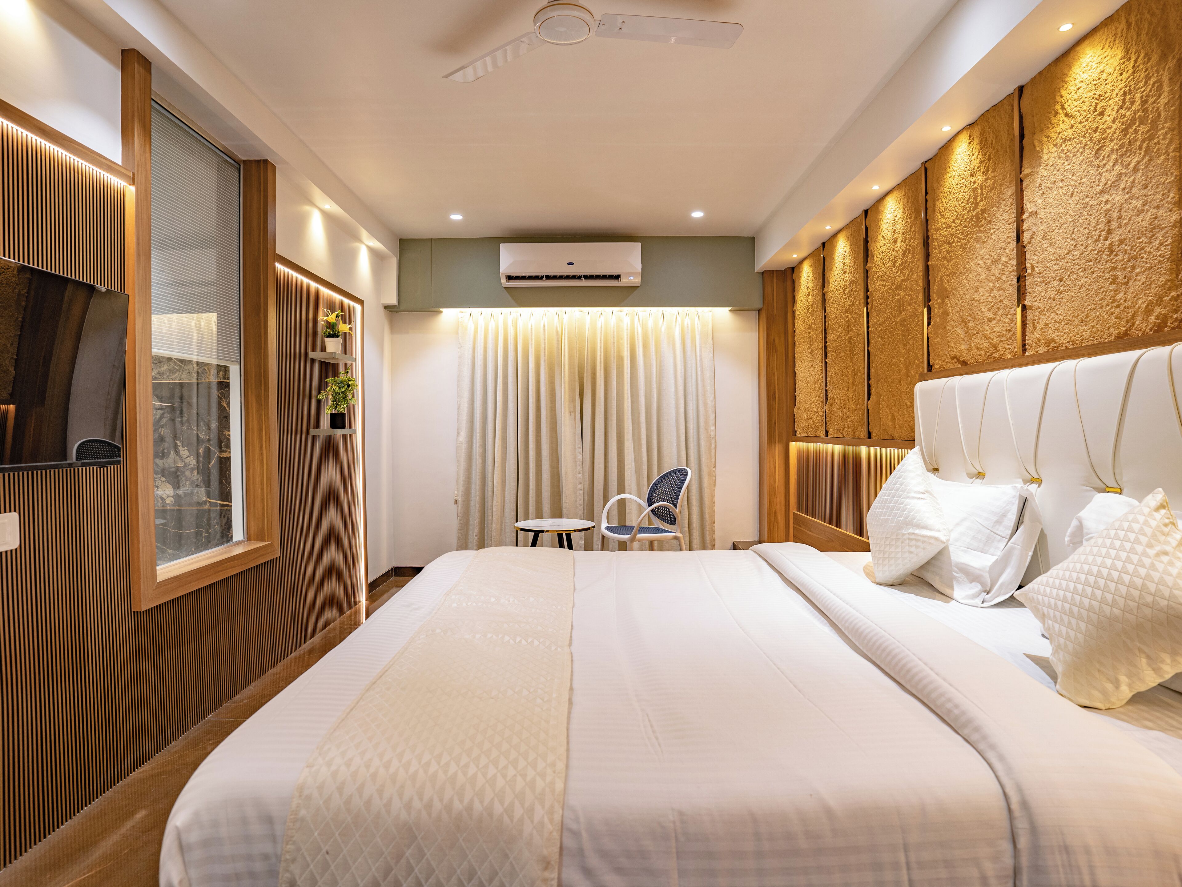A modern hotel room with a large bed, a ceiling fan, an air conditioning unit, a window with blinds, a small table, and a chair.