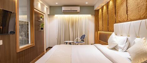 A modern hotel room with a large bed, a ceiling fan, an air conditioning unit, a window with blinds, a small table, and a chair.