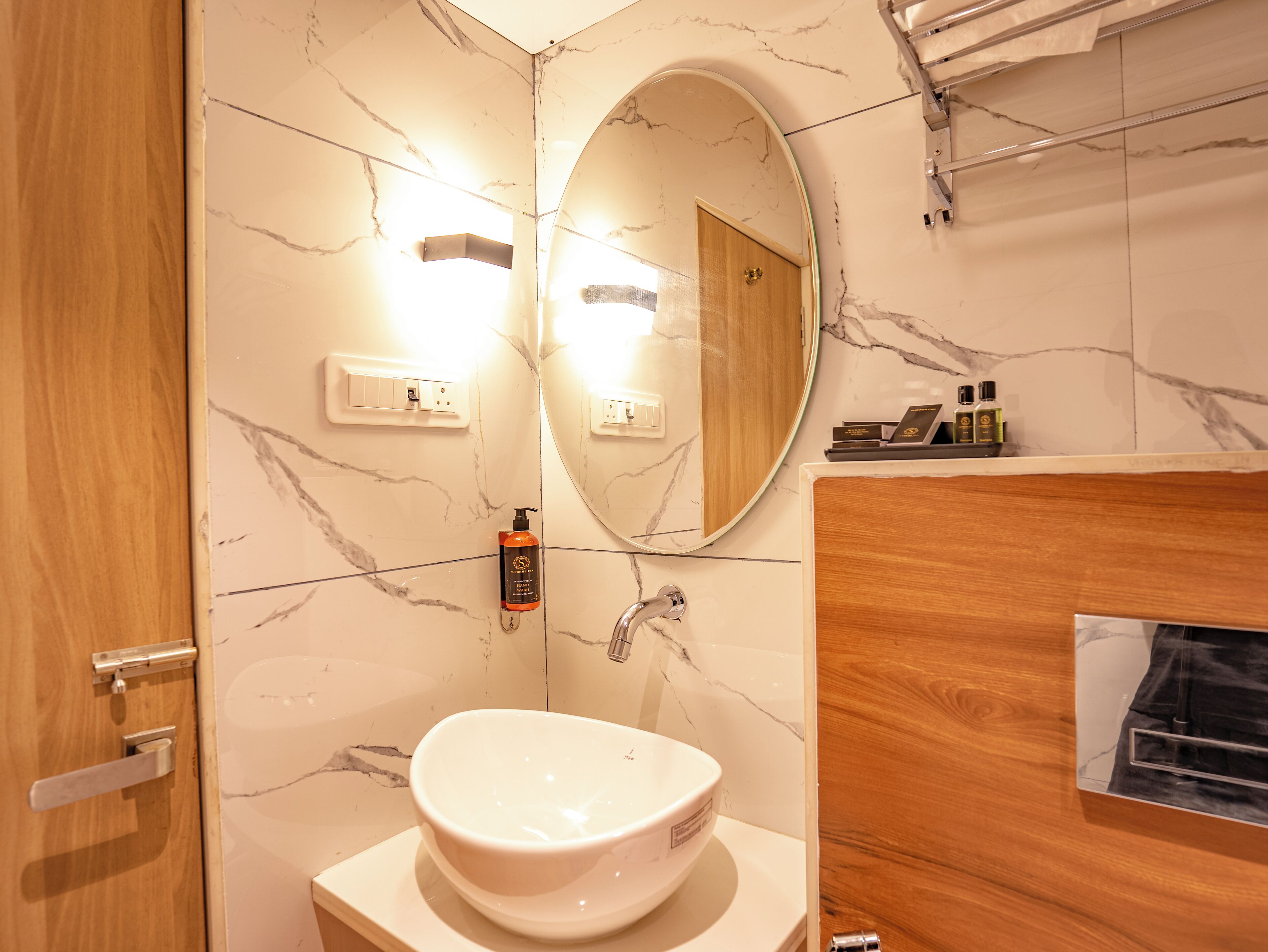 A bathroom with a marble wall, a wooden door, a white sink, a mirror, and a vanity with toiletries.