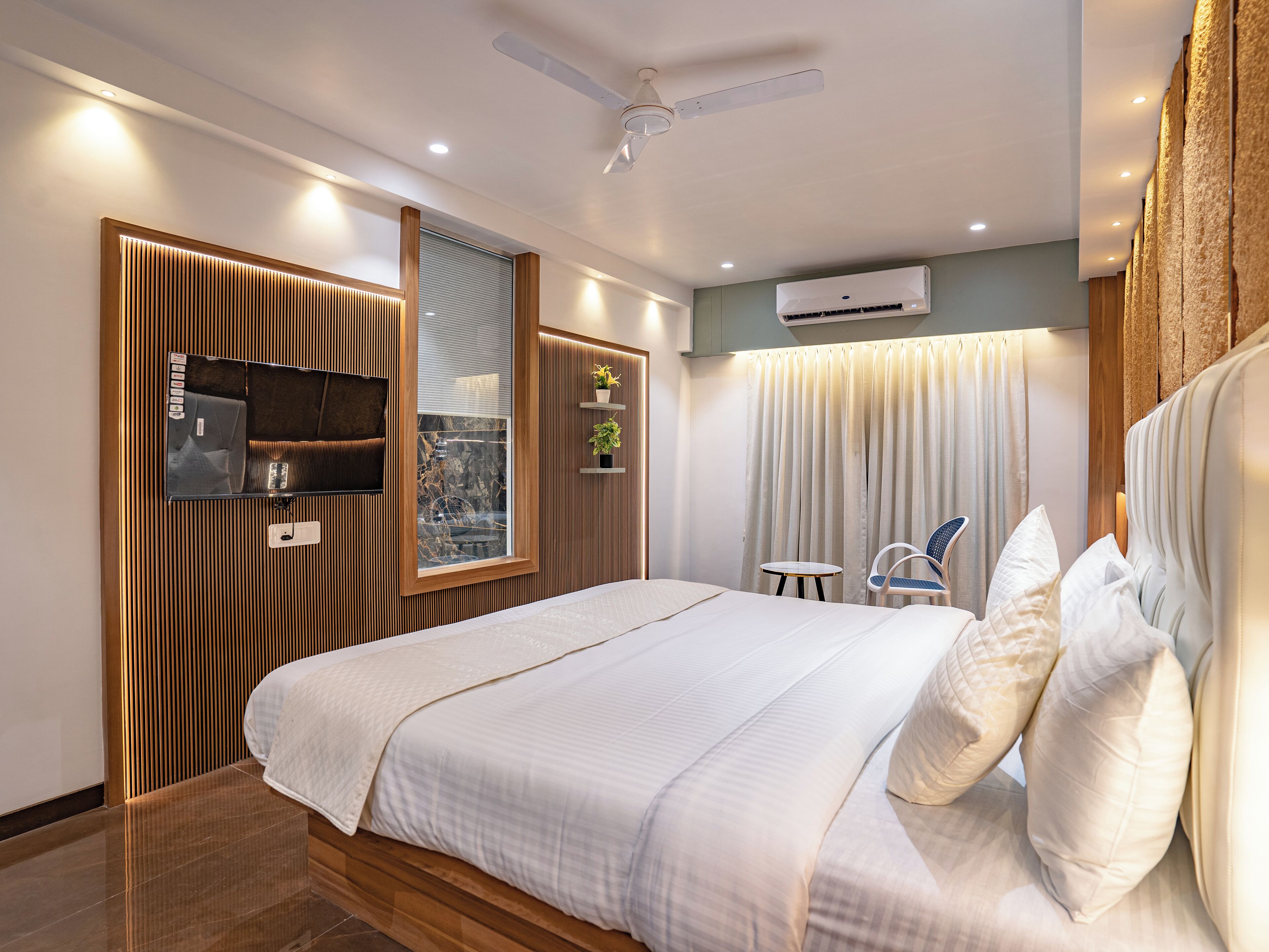 A modern bedroom with a large bed, a TV mounted on the wall, a ceiling fan, and an air conditioning unit.
